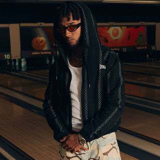 En mand står på en bowlingbane iført en sort hættetrøje med mønstrede syninger, hvid tanktop og lyse camouflageshorts. Han bærer solbriller og har hætten oppe, hvilket giver et cool, afslappet look i den stemningsfulde bowlinghal.