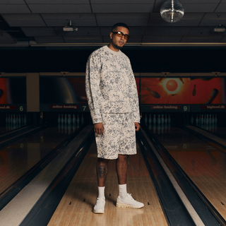 En mand står midt i en bowlingbane iført et matchende sæt med langærmet trøje og shorts i lysegråt camouflagemønster. Han har hvide sokker, beige sneakers og solbriller på, og poserer selvsikkert under en diskokugle i det dæmpede lys.