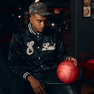 En mand sidder i en bowlinghal iført en sort satin varsity-jakke med teksten "E Last" i hvid skrift og en camouflagekasket. Han kigger ned mod en rød bowlingkugle, han hviler hånden på, i en afslappet og eftertænksom positur.